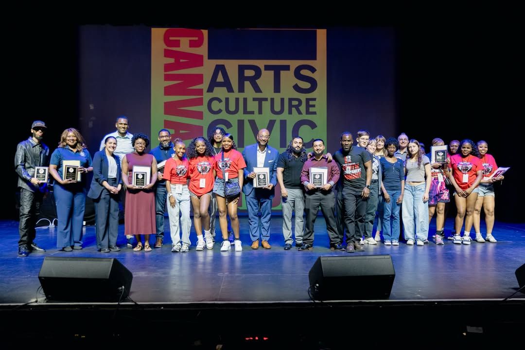 A large group of adults and young people standing together on a theater stage, with several people holding recognition plaques.